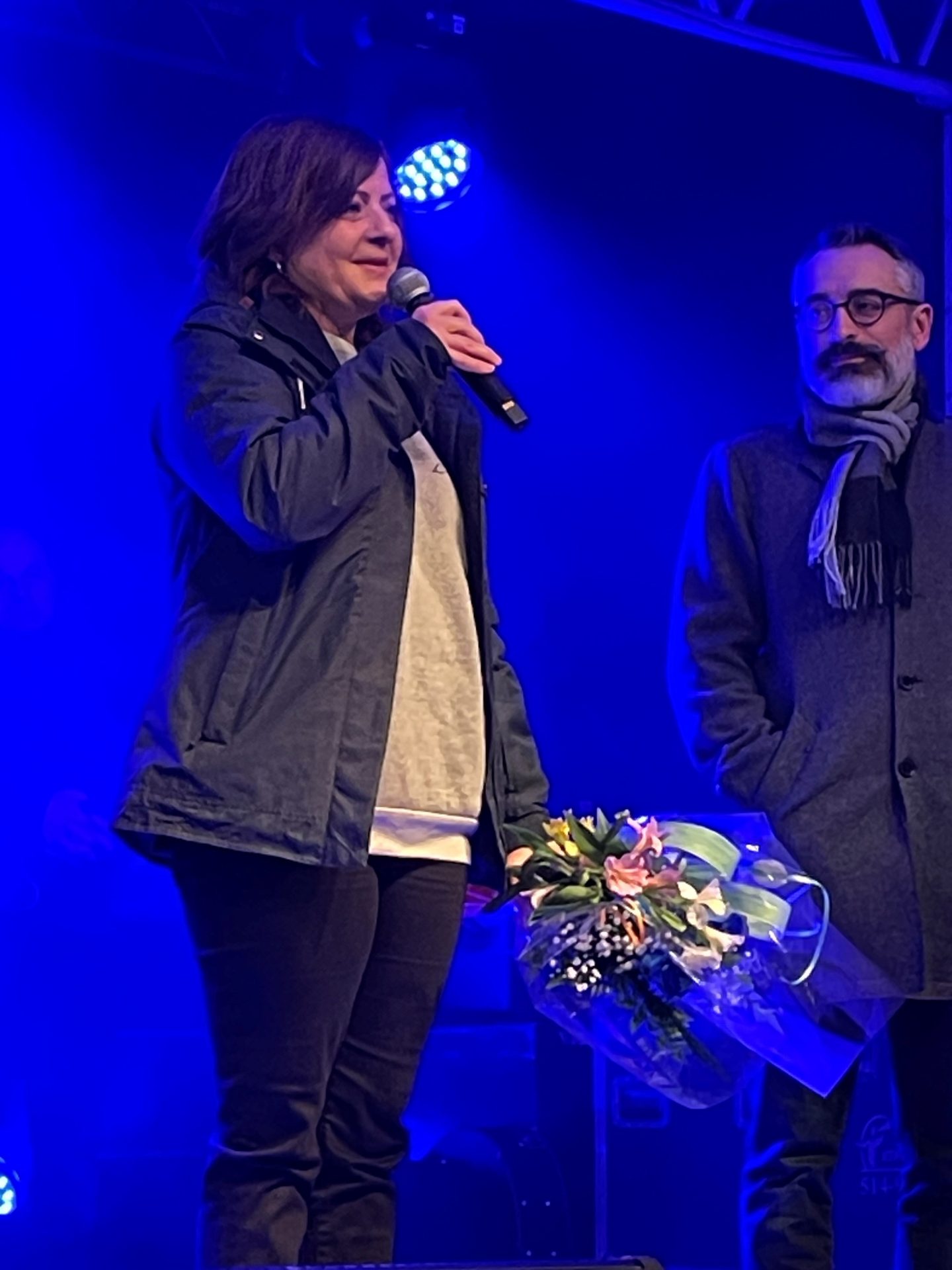 Une femme tient un bouquet de fleurs sur scène et parle au micro.